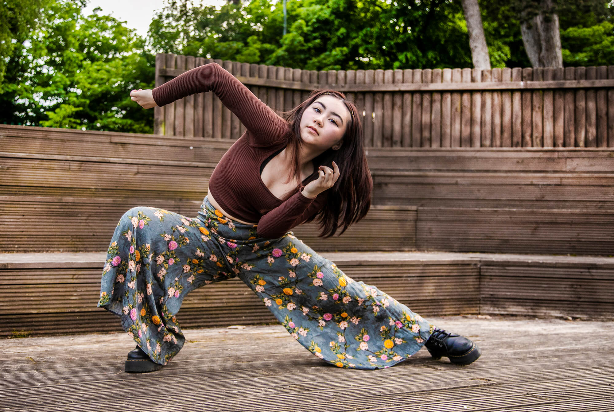 global majority female dancer lunging to the left with albow pointed in the air. In front of soft focus trees and wooden gate. Wearing brown long sleeved t-shirt and flowered flared trousers. 