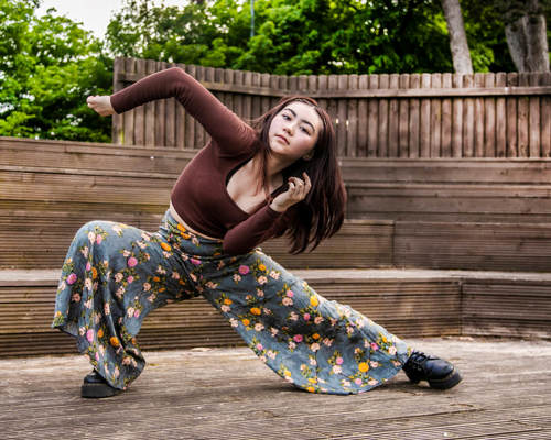 global majority female dancer lunging to the left with albow pointed in the air. In front of soft focus trees and wooden gate. Wearing brown long sleeved t-shirt and flowered flared trousers. 