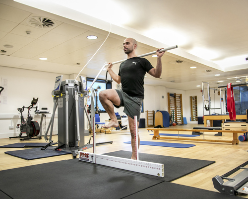 A bald male standing on one leg stepping over floor ruler, holding a pole behind his back. In a gym with lots of excersise equpiment in the background 