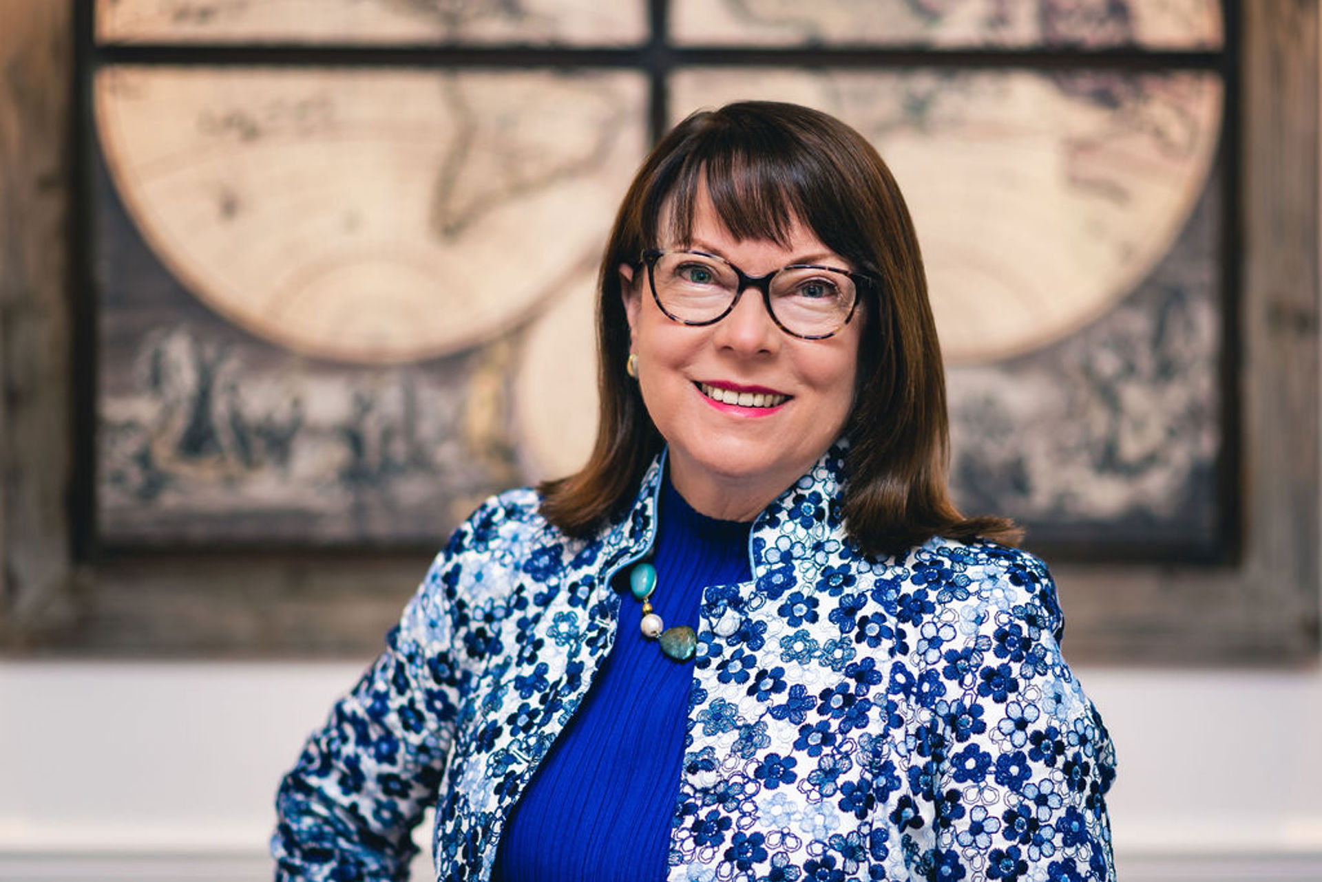 white female with brown hair and rectangle glasses. Wearing blue floral blazer and blue top