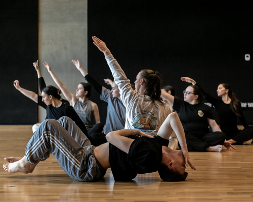7 female dancers sitting crossed legged with one arm in the air. one male dancer infront laying on the floor with knees up arched back and hand infront of face. In a dance stucio with wooden floors and black walls 