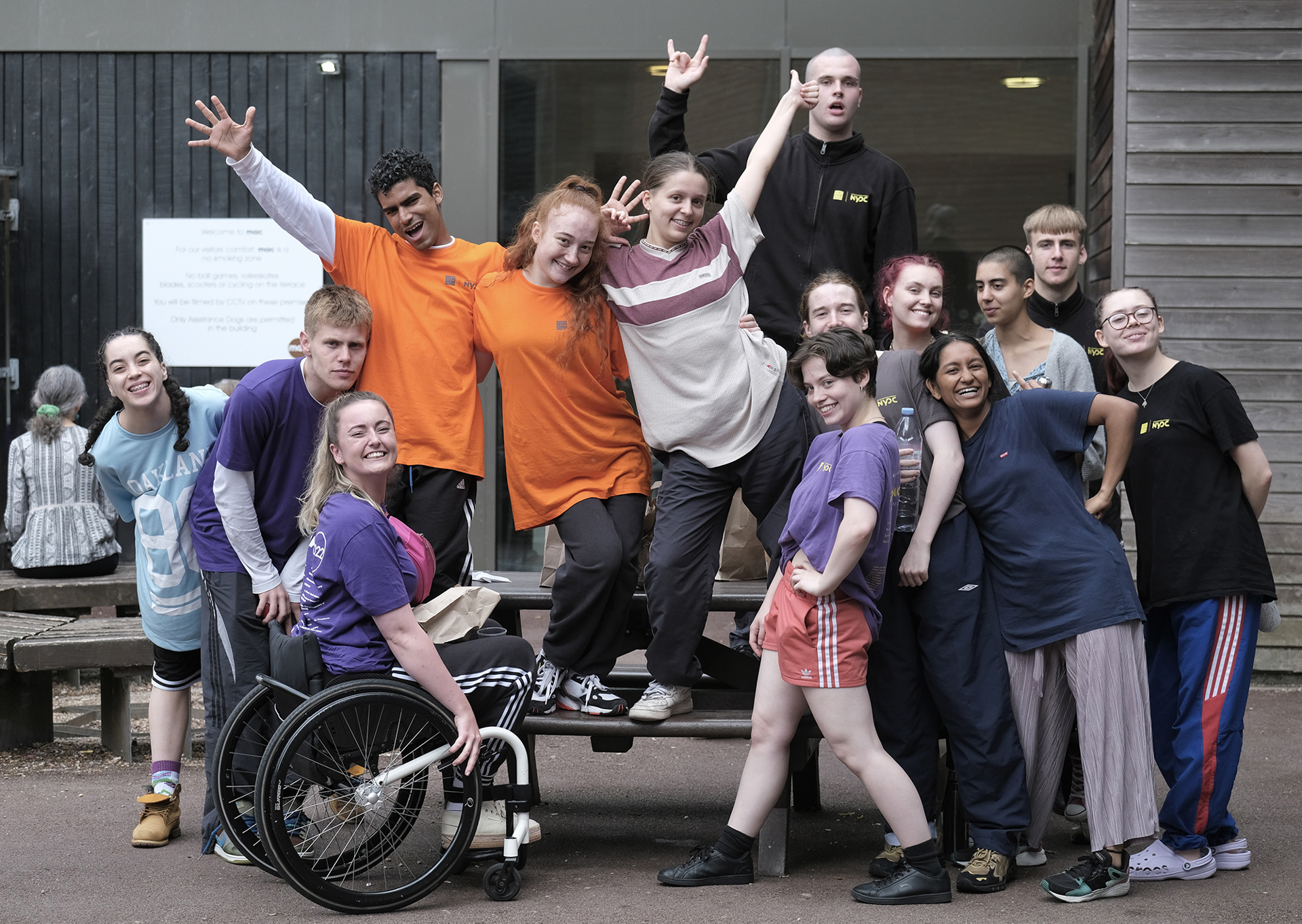 Group of mixed male and female, different races, differently abled young people posing for a picture some sitting on a bench others arms up standing iwth one wheel chair dancer.