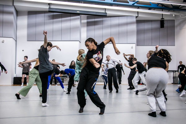Young people dancing in a dance studio all interacting with each other. Main focus on global majority female dancer moving her arms to the right and swinging hair around.