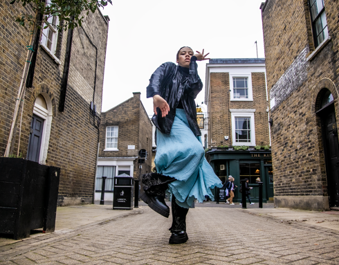 Global majority female dancer dancing down london street facing camera one arm near the face and on one leg . Wearing black leather jacket and blue skirt