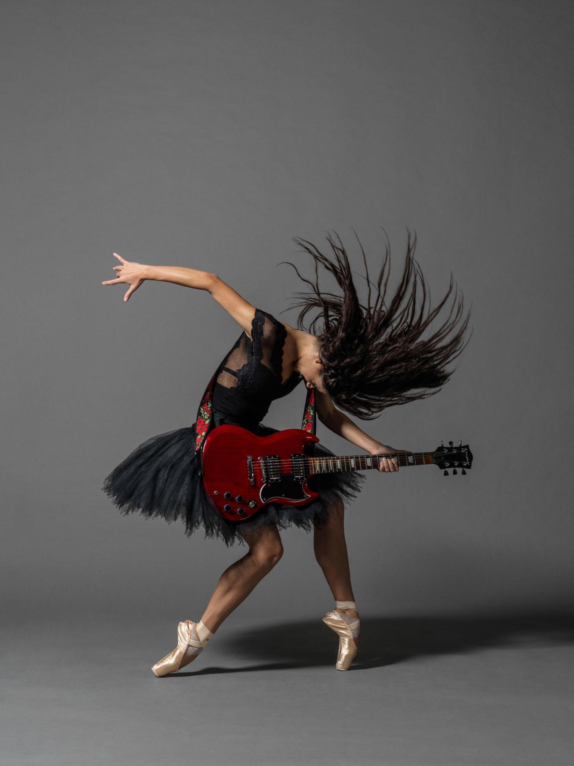 female dancer on point with bended knees, holding a guitar arching over it, with one hand flung behind and long brown hair flicked over the head