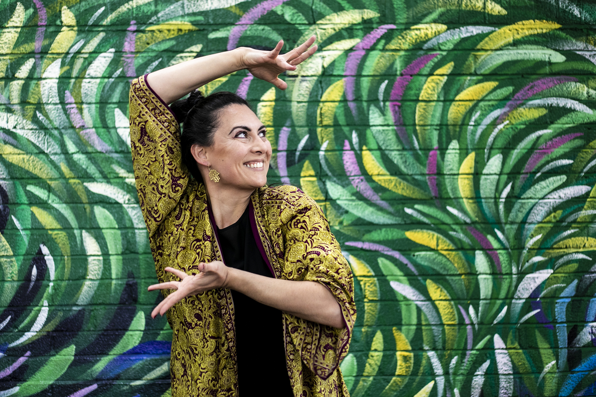 Global majority female dancer with long black hair smiling with arms framing the face. In front of green colourful graffiti wall. Wearing green and purple kaftan. Global majority female dancer with long black hair smiling with arms framing the face. In front of green colourful graffiti wall. Wearing green and purple kaftan.