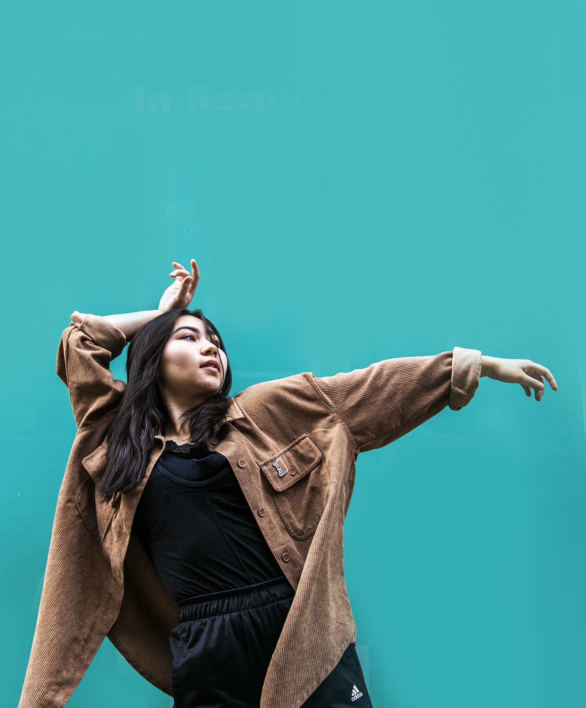 Young female global majority dancer with one arm rested on head and other pointed to the right, with head facing top right of the screen. In front of blue background, wearing brown shirt and black trousers. 