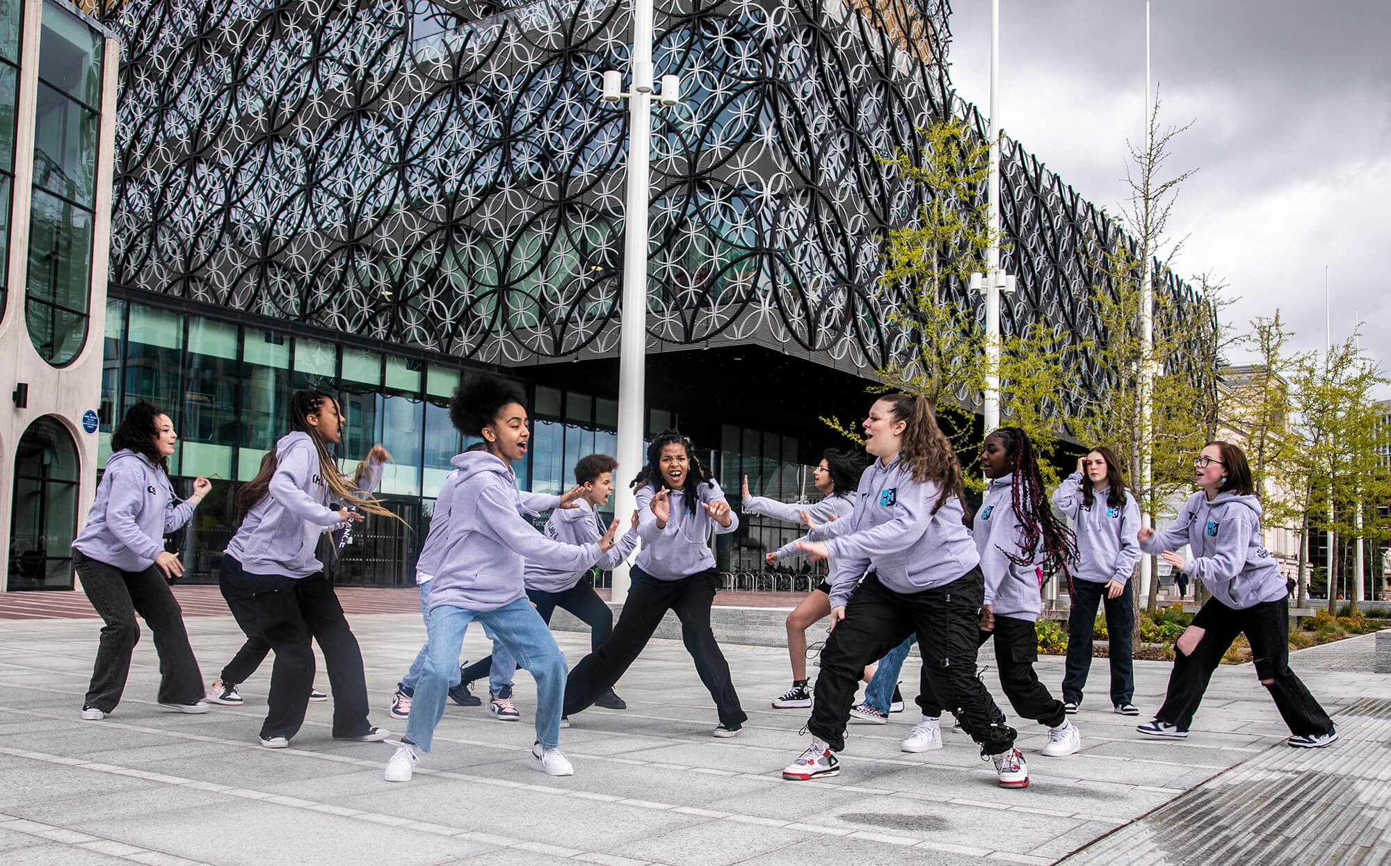 Group of teenage female dancers performing street dance infront of large building with circles. All wearing matching grey hoodies 