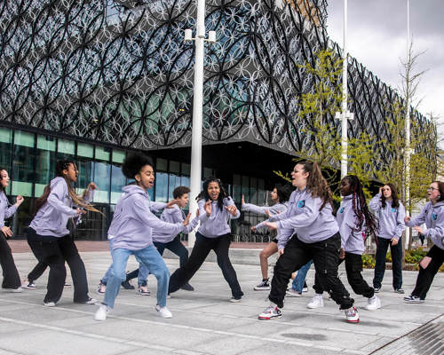 Group of teenage female dancers performing street dance infront of large building with circles. All wearing matching grey hoodies 