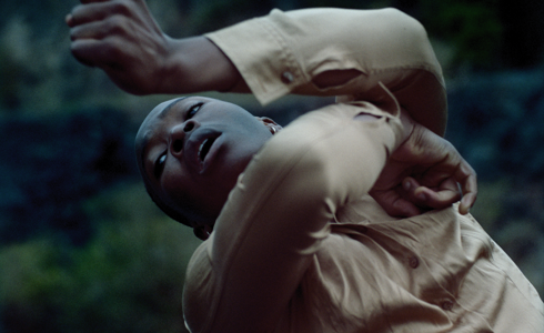 Close up of global majority dancer moving arms around the face with eyes up to the top left corner of the image, wearing muted clothes in front of dark wall 