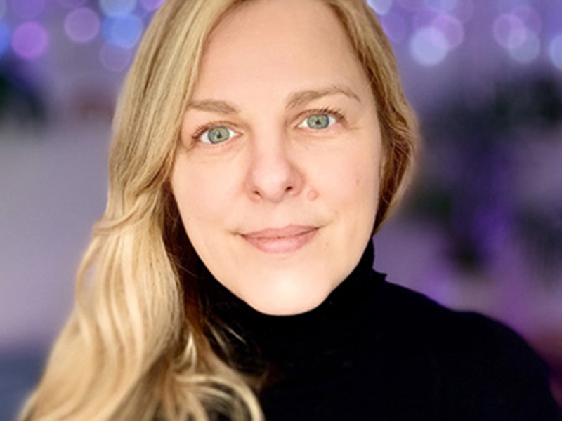 Headshot of Sarah Pritchard. White blonde female looking at the camera wearing high neck black top with purple background