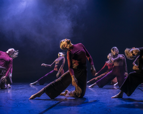 6 female dancers performing on the U.Dance stage wearing long sleve red tops and baggy brown trousers. All staners on one know with the other leg straight to the left with heads titled to the sky. Stage lighting is dark with blue fog and floor. 