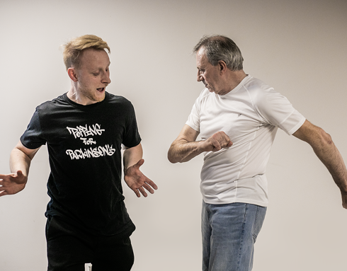 Two men body poppin dancing together. One young blonde white male dance teacher wearing all black, the other an older male grey haired dance student wearing blue jeans and white top. Two men body poppin dancing together. One young blonde white male dance teacher wearing all black, the other an older male grey haired dance student wearing blue jeans and white top.