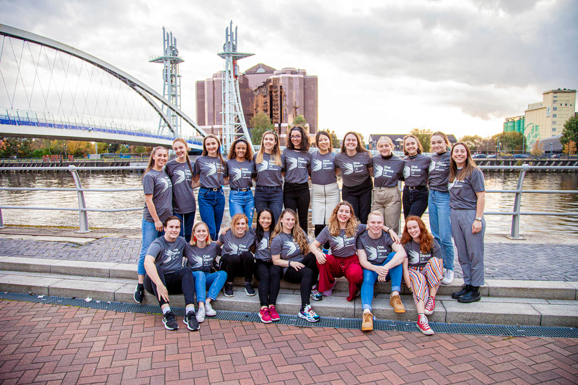 20 male and female dance ambassadors linked arms smiling in front of Manchester bridge and skyline in the background. Wearing grey Dance ambassador tshirts. 20 male and female dance ambassadors linked arms smiling in front of Manchester bridge and skyline in the background. Wearing grey Dance ambassador tshirts.