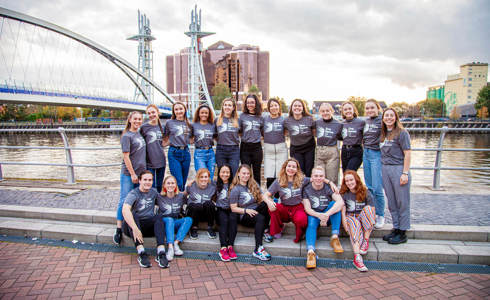 20 male and female dance ambassadors linked arms smiling in front of Manchester bridge and skyline in the background. Wearing grey Dance ambassador tshirts.