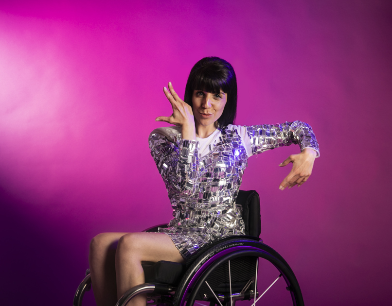 white brunette female dancer in a wheelchair wearing silver sparkly dress. Both hands in salsa movement around her face. On a pink photo backdrop