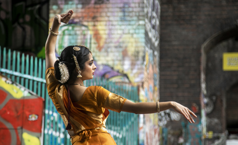 Back of a South Asian dancer dressed in traditional orange dress with hair jewellery. In front of colourful graffiti walls. One arm above her head and one arm stretched out. 
