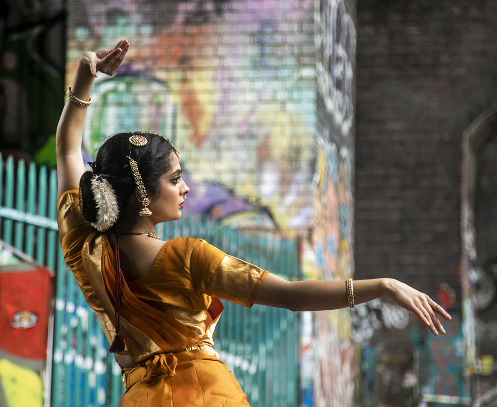 Back of a South Asian dancer dressed in traditional orange dress with hair jewellery. In front of colourful graffiti walls. One arm above her head and one arm stretched out.