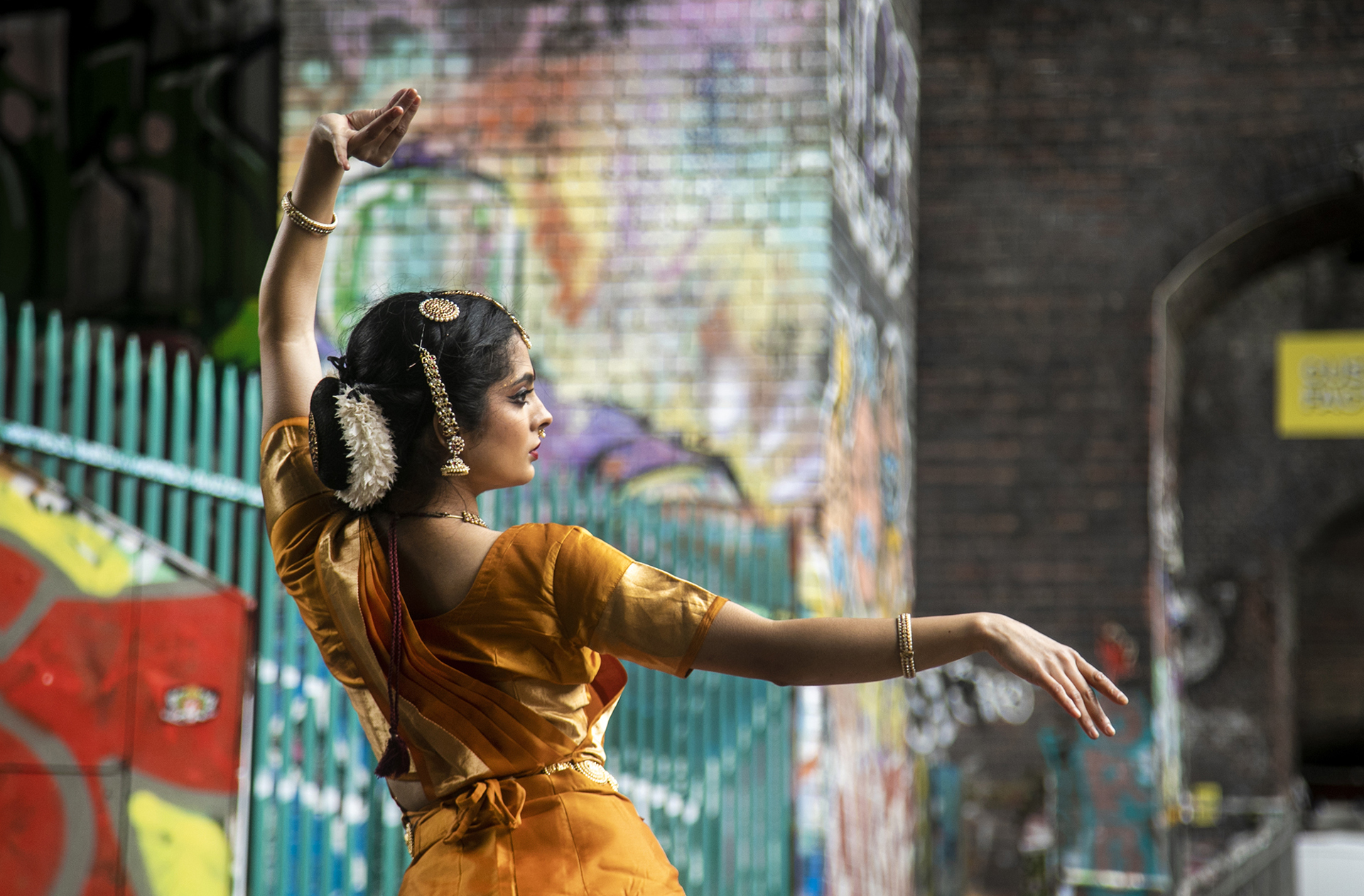 Back of a South Asian dancer dressed in traditional orange dress with hair jewellery. In front of colourful graffiti walls. One arm above her head and one arm stretched out. Back of a South Asian dancer dressed in traditional orange dress with hair jewellery. In front of colourful graffiti walls. One arm above her head and one arm stretched out.