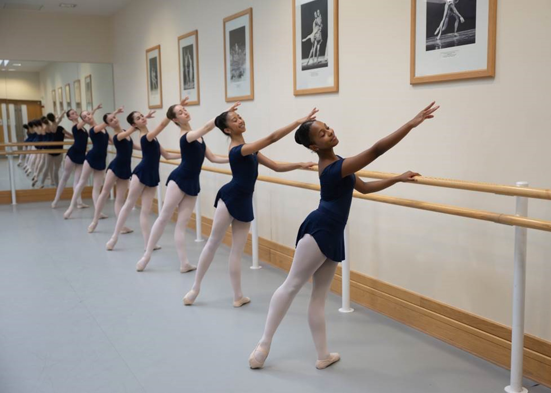 severn female young ballet dancers holding on to a ballet bar with one arm pointed to the top left of the image and the bottom left foot pointed away. Wearing blue leotards and pink tights.