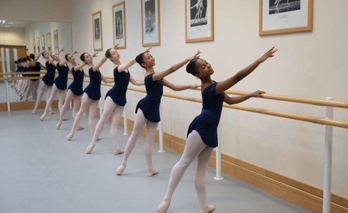 severn female young ballet dancers holding on to a ballet bar with one arm pointed to the top left of the image and the bottom left foot pointed away. Wearing blue leotards and pink tights.