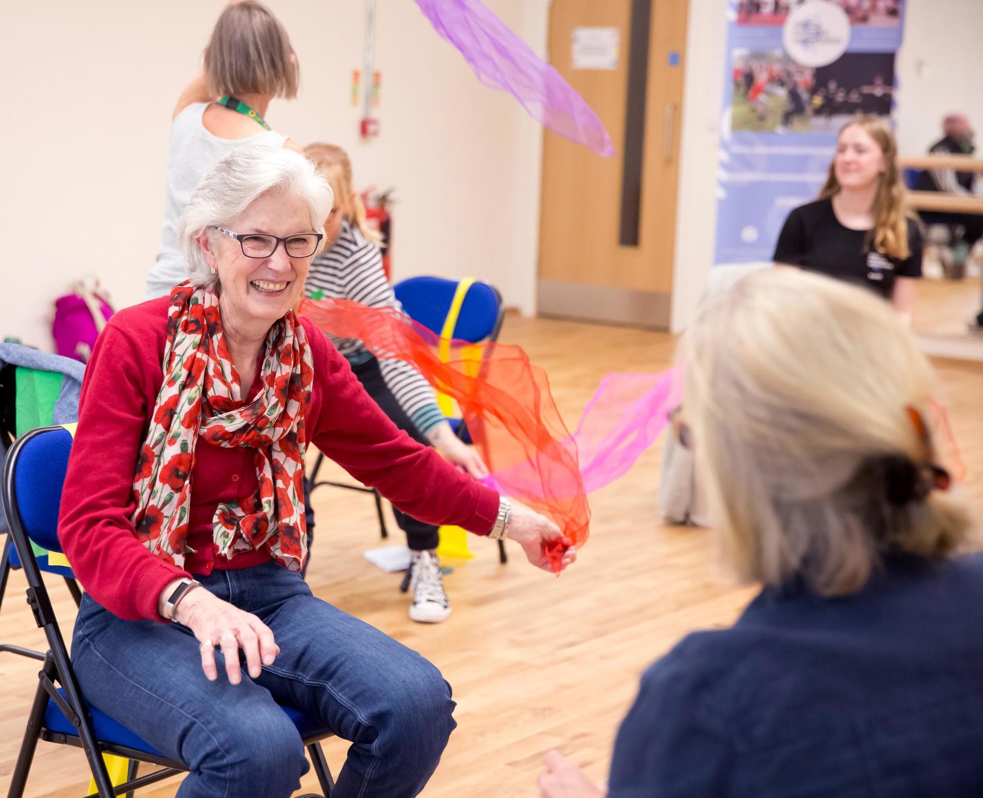 Older female sitting on a chair waving a colourful scarf around whilst smiling Older female sitting on a chair waving a colourful scarf around whilst smiling