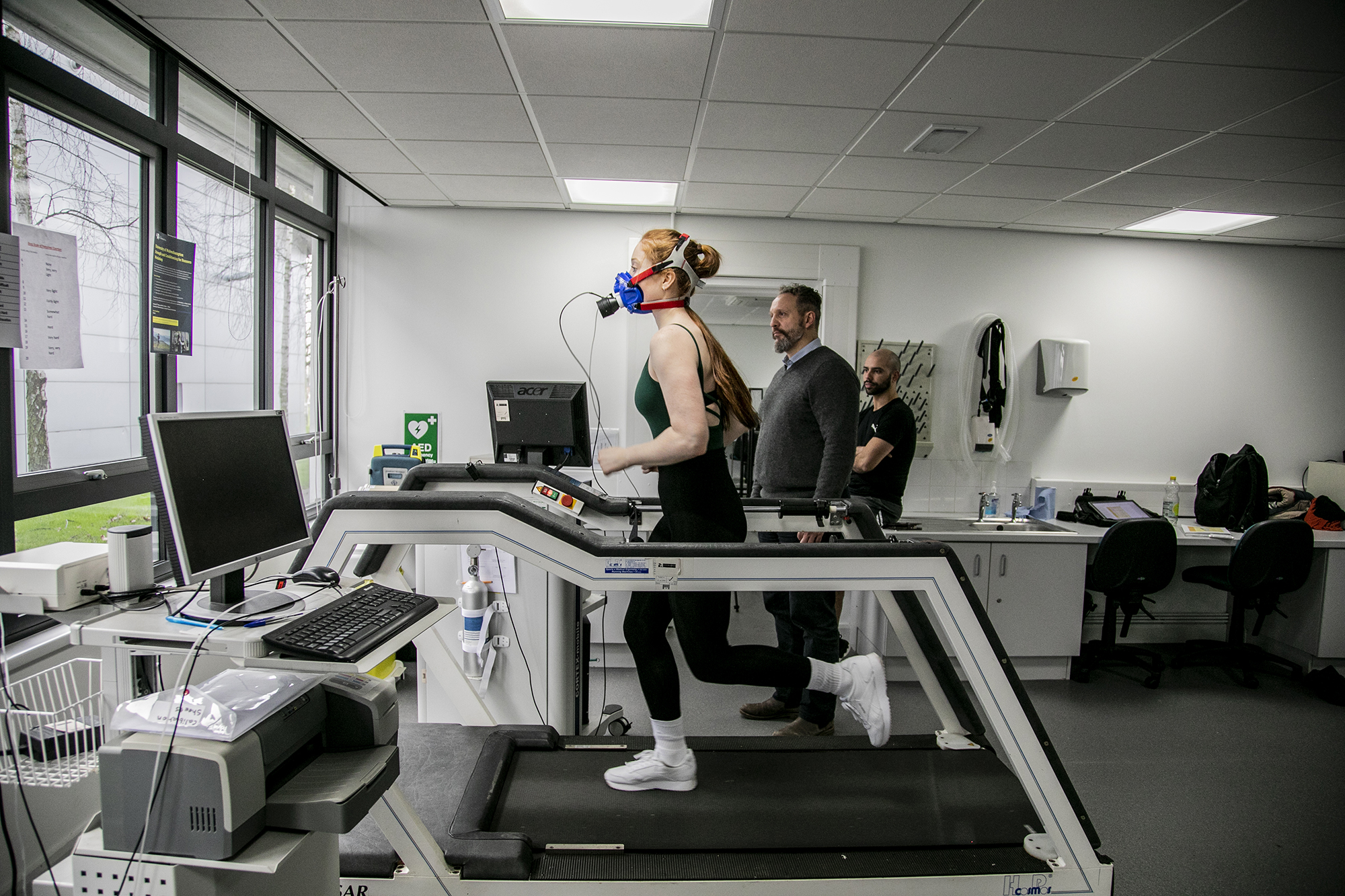 White female dancer with long red hair running on a treadmill wearing face-mask to measure cardiovascular fitness. White male physiologist looking over to observe stats on the machine. In a white laboratory setting with a window in the background