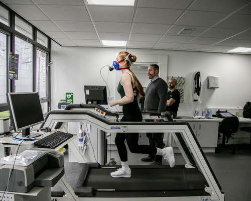 White female dancer with long red hair running on a treadmill wearing face-mask to measure cardiovascular fitness. White male physiologist looking over to observe stats on the machine. In a white laboratory setting with a window in the background