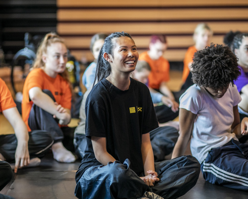 young dancers white and global majority, male and female, sitting crossed legged smiling and listening. 