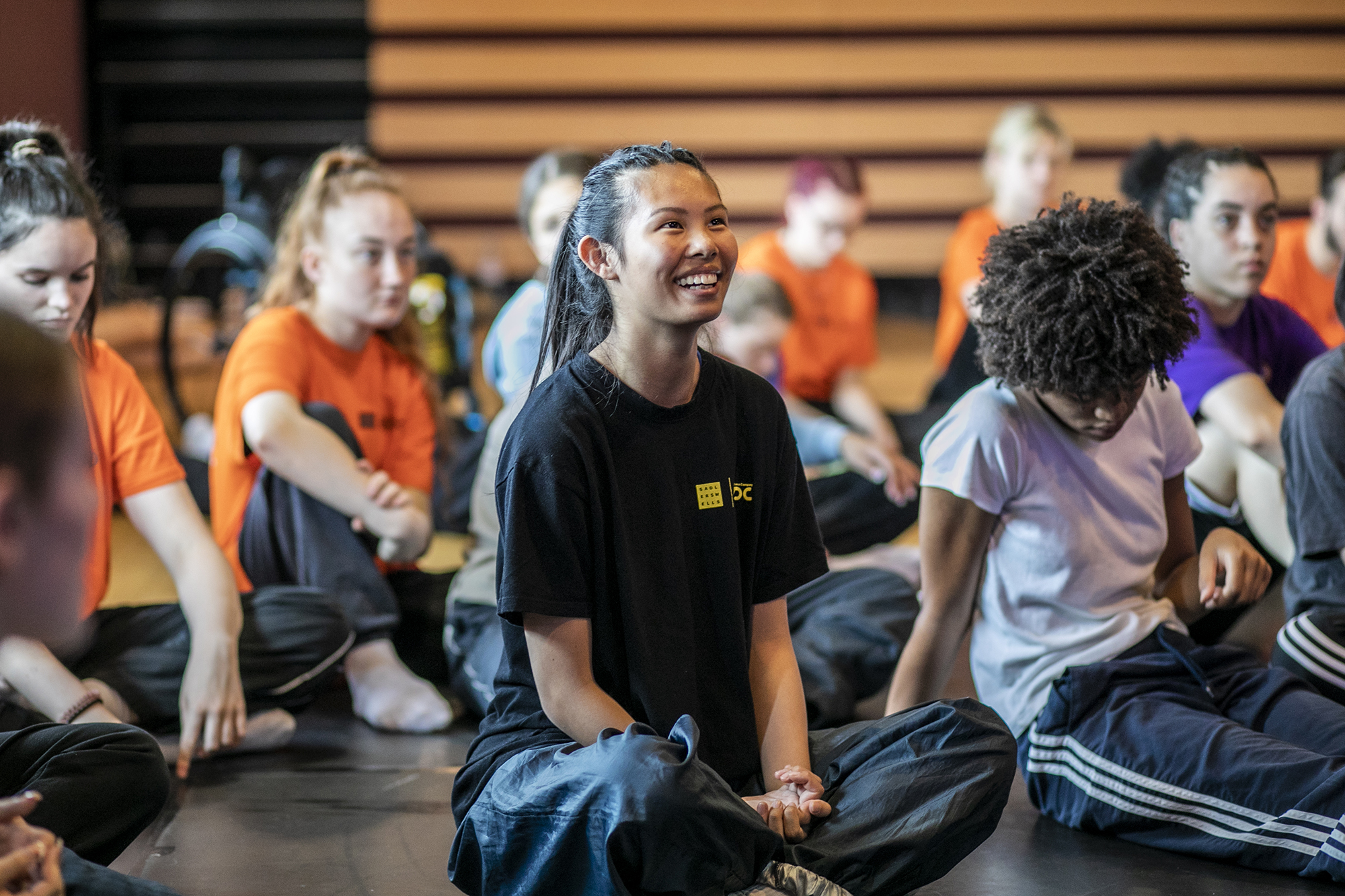 young dancers white and global majority, male and female, sitting crossed legged smiling and listening.  young dancers white and global majority, male and female, sitting crossed legged smiling and listening.