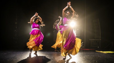 Three female Bollywood dancers spinning on a dark stage wearing pink and yellow sari dresses.