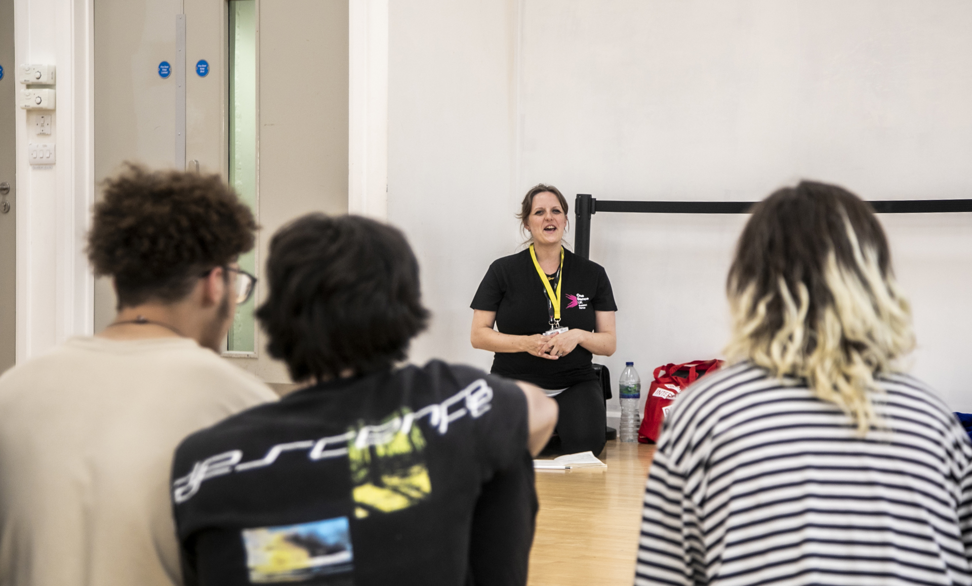 One Dance UK staff member sitting on knees talking to three students sitting in front of the camera in a dance studio. One Dance UK staff member sitting on knees talking to three students sitting in front of the camera in a dance studio.