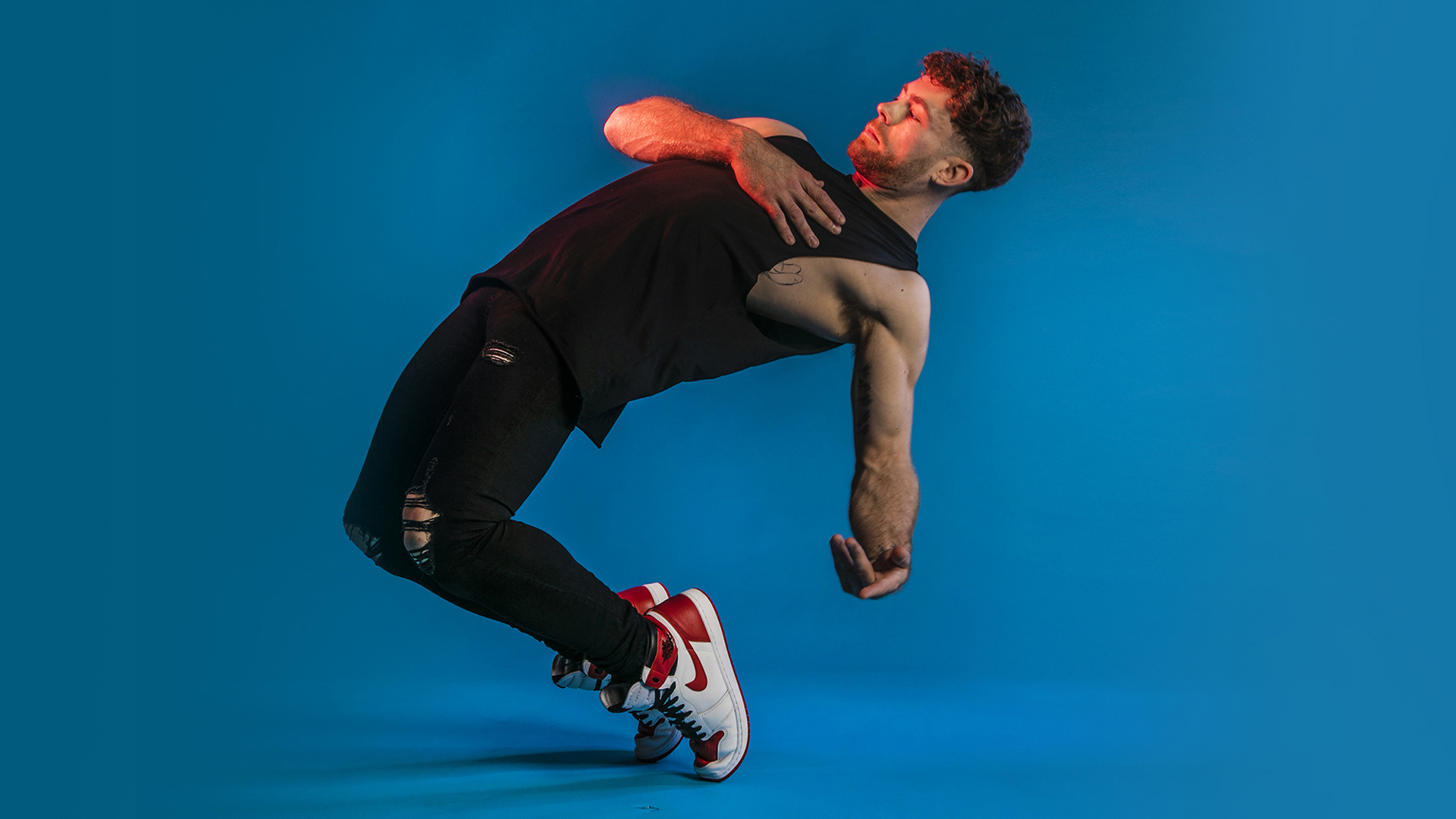 white male dancer leaning back on his tip toes. Black outfit and white trainers. On blue studio background. 