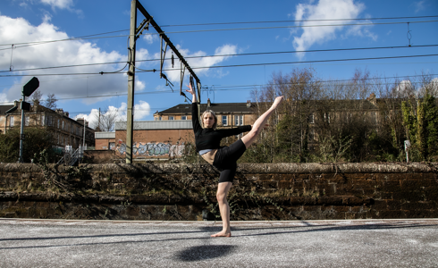 white female cropped blonde hair on a train platform with one leg kicked high to the side up, another on demi point and one arm above and another to the right. Wearing black shorts and black crop top with bare feet 