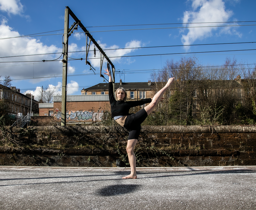 white female cropped blonde hair on a train platform with one leg kicked high to the side up, another on demi point and one arm above and another to the right. Wearing black shorts and black crop top with bare feet