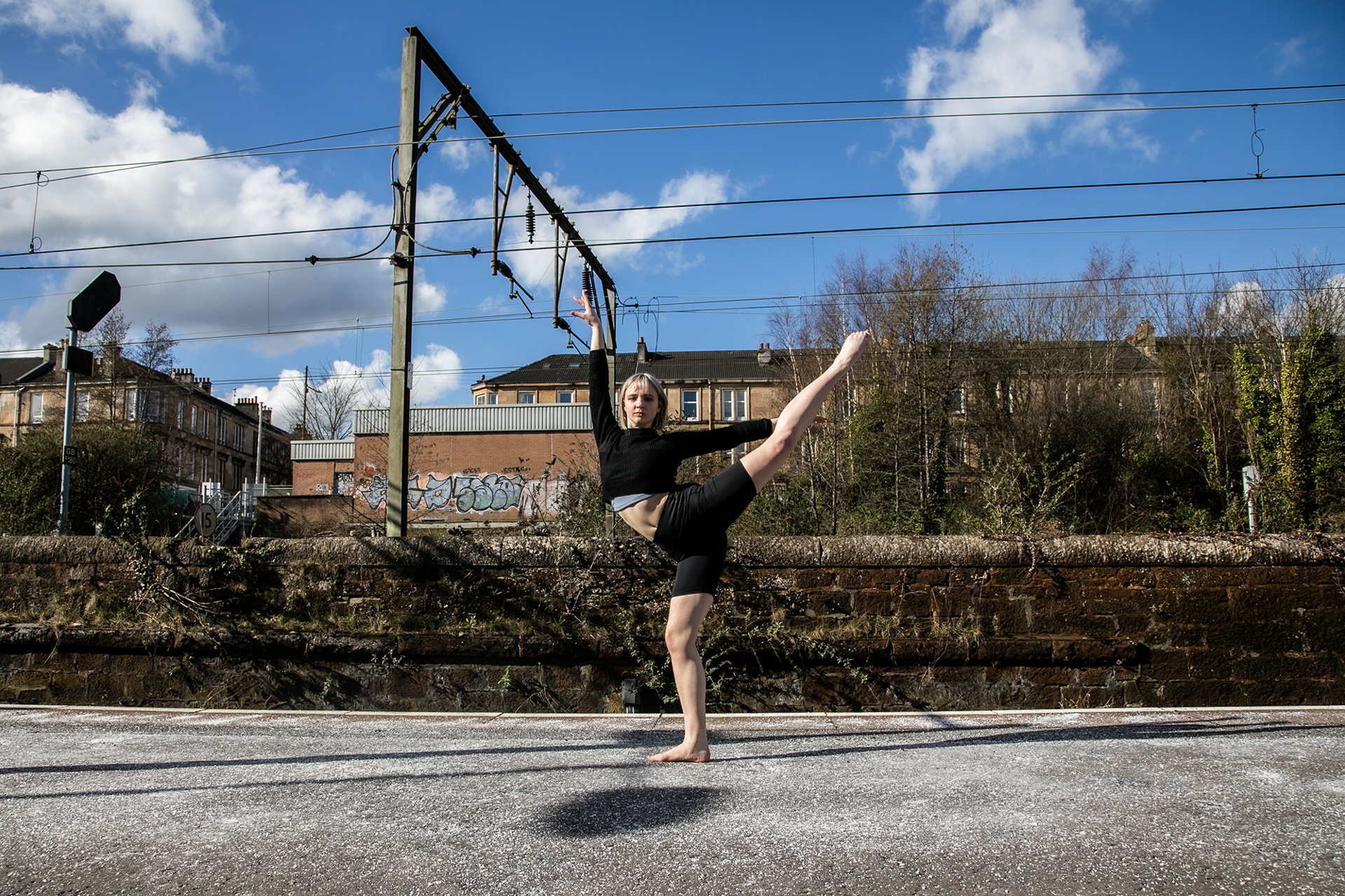 white female cropped blonde hair on a train platform with one leg kicked high to the side up, another on demi point and one arm above and another to the right. Wearing black shorts and black crop top with bare feet 