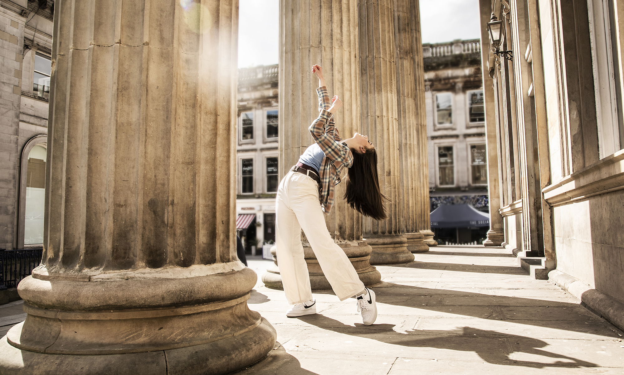 Young female dancer infront of Glasgow coucil building with large pillars. Legs lunched arms above the head. White female dance with long brown hair wearing checked shirt blue top and white trousers  Young female dancer infront of Glasgow coucil building with large pillars. Legs lunched arms above the head. White female dance with long brown hair wearing checked shirt blue top and white trousers
