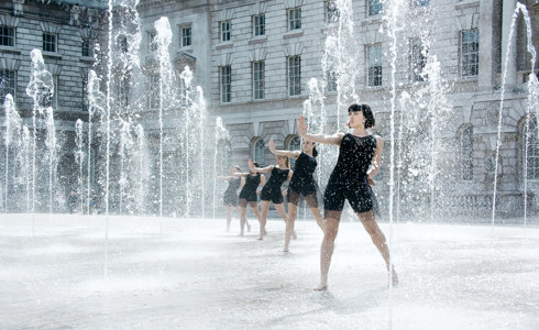 five female dancers with one arm reached out all standing in a water fountain. Wearing black outfits.