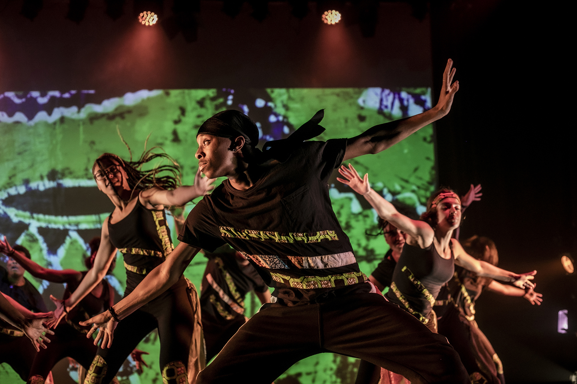 young people performing on udance stage. Main focus is on black male dancer arms stretched diagonally and looking to the left wearing black with camo striped and a black durag. Backdrop is green patterns with red lights 