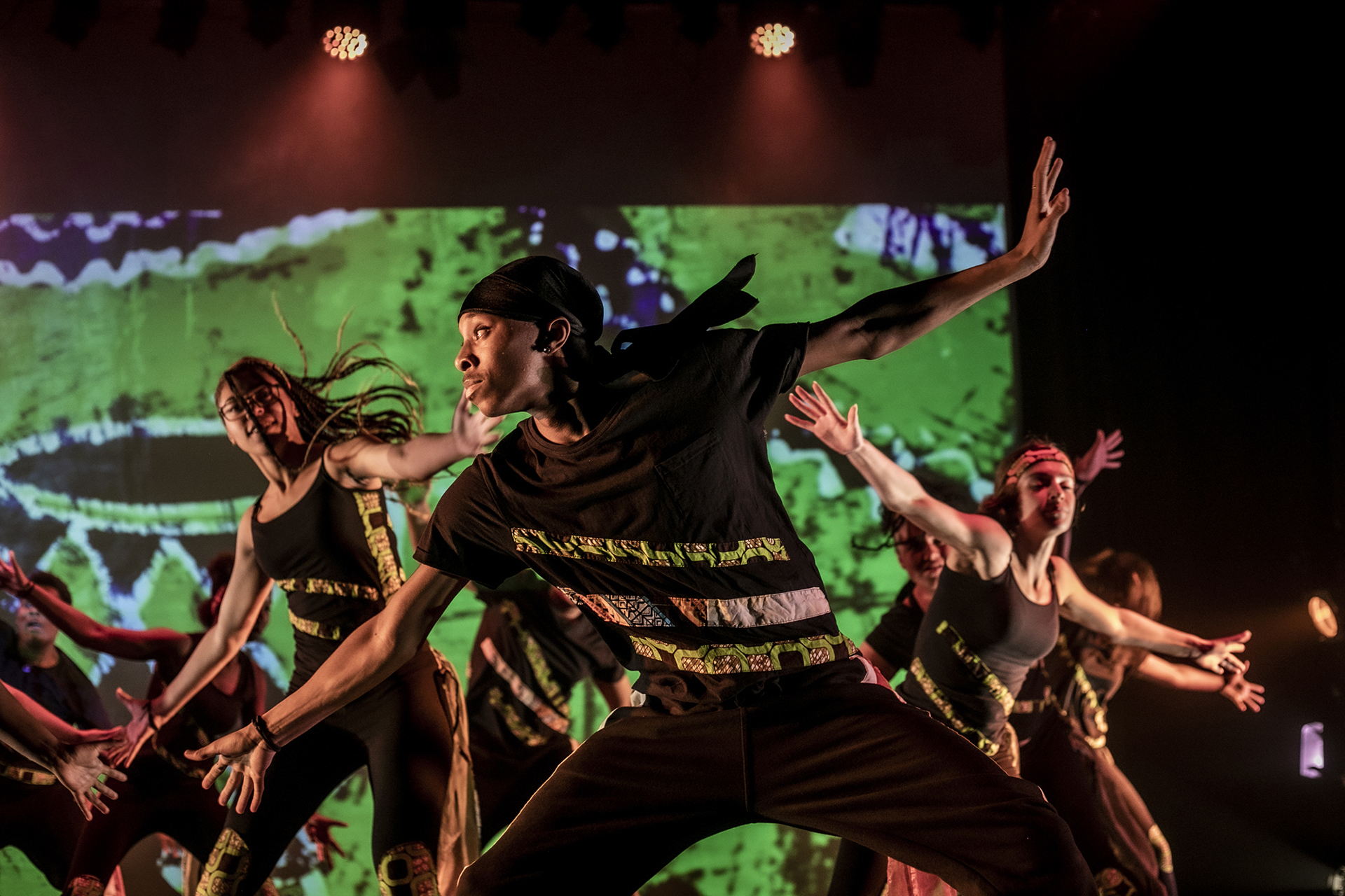 young people performing on udance stage. Main focus is on black male dancer arms stretched diagonally and looking to the left wearing black with camo striped and a black durag. Backdrop is green patterns with red lights young people performing on udance stage. Main focus is on black male dancer arms stretched diagonally and looking to the left wearing black with camo striped and a black durag. Backdrop is green patterns with red lights