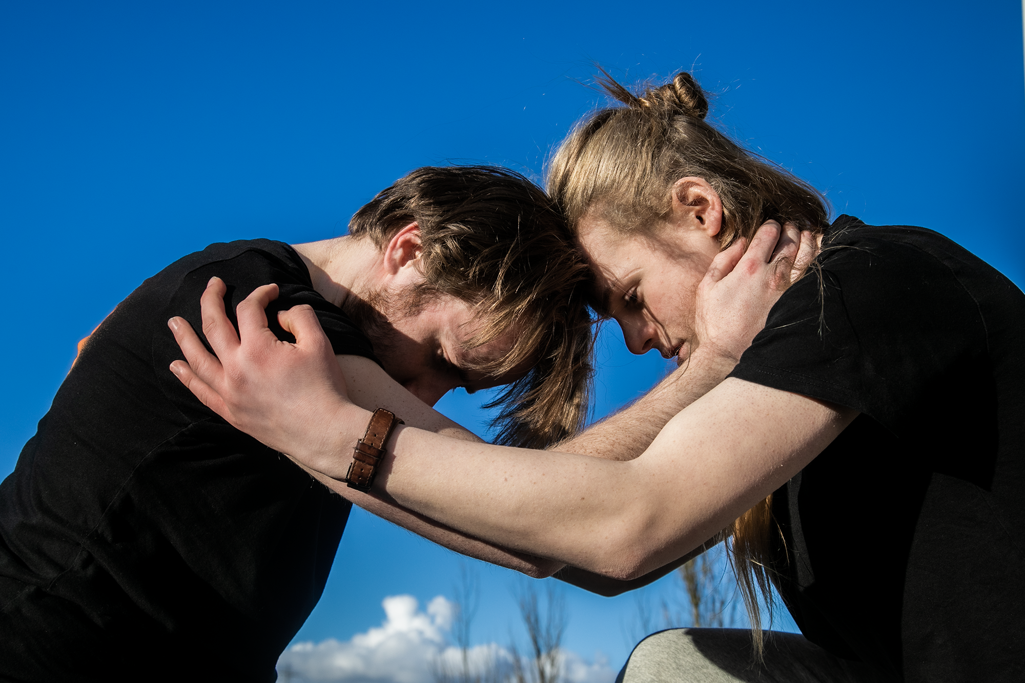 Two white male dancers holding eachothers forearms with heads together looking down with blue sky background.