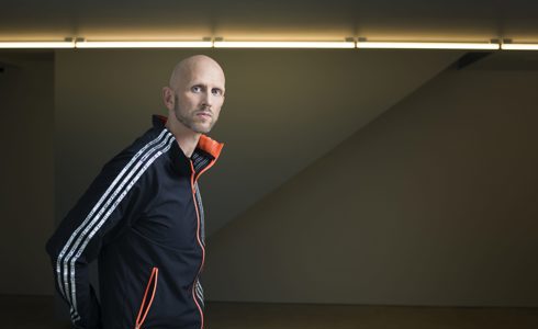 headshot of Wayne Mcgregor. White bald male with stubble standing to the left of image wearing a blue tracksuit zip up 