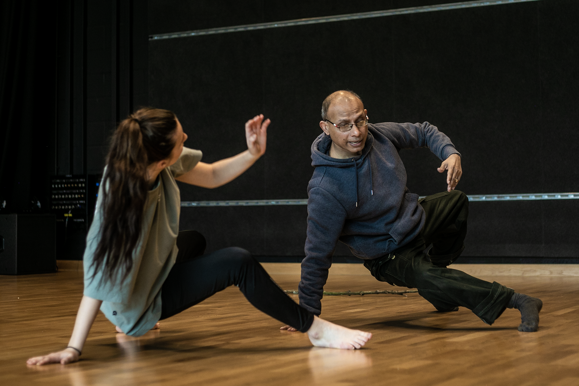 older male global majority dancer teaching white young female dancer. Both on the floor leaning on one hand.