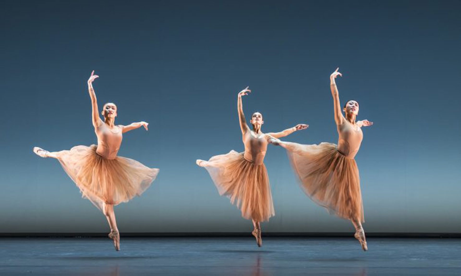 Three female ballet dancers wearing nude leotards and long tutus in a jumping arabesque with both arms high on blue stage