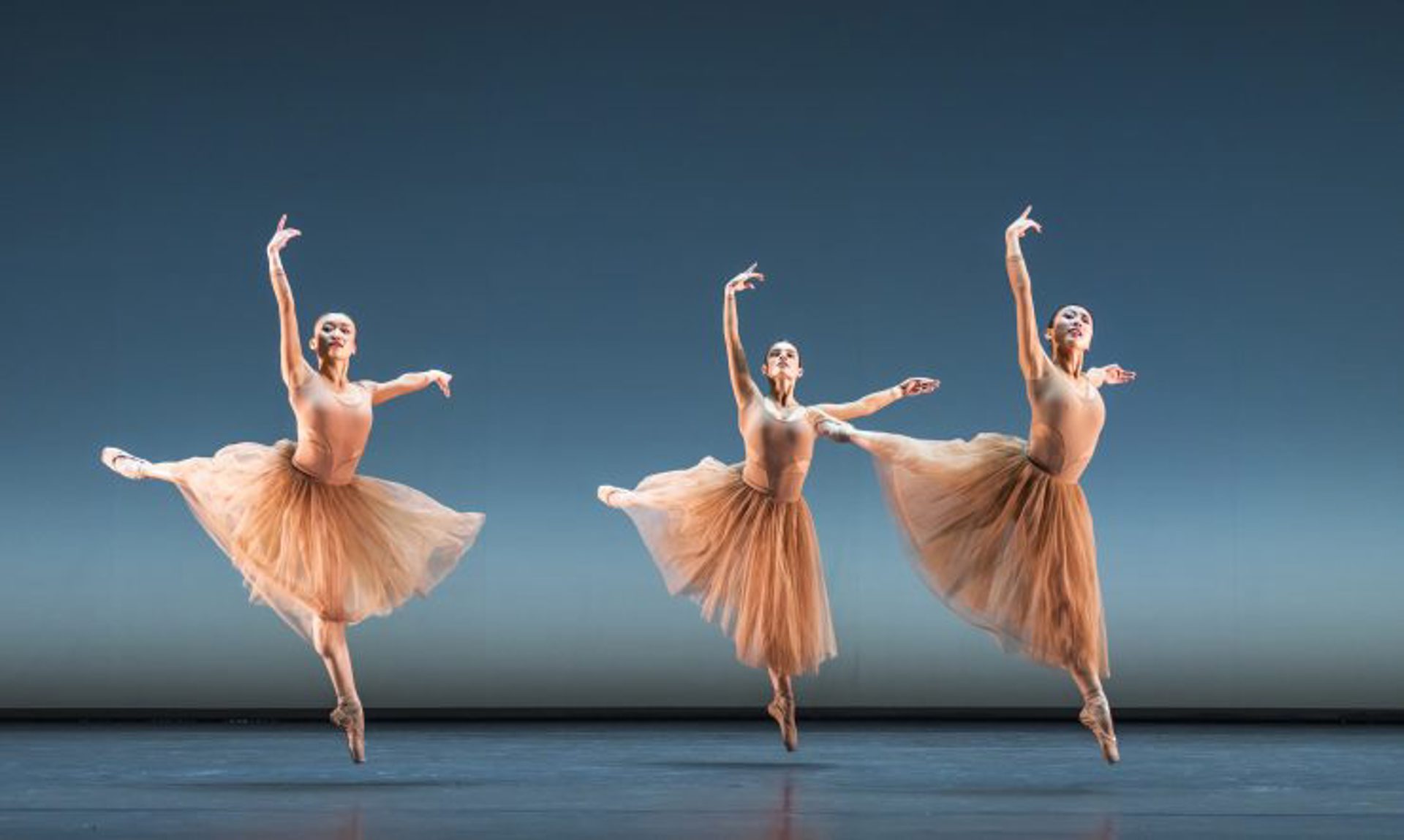 Three female ballet dancers wearing nude leotards and long tutus in a jumping arabesque with both arms high on blue stage Three female ballet dancers wearing nude leotards and long tutus in a jumping arabesque with both arms high on blue stage