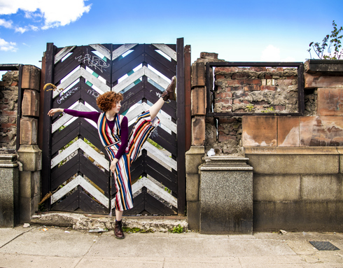 white female dancer with red short curly hair and striped jumpsuit, high kicking right leg with left hand to the side. In front of triangle painted black and white gate with brick wall and blue sky above.