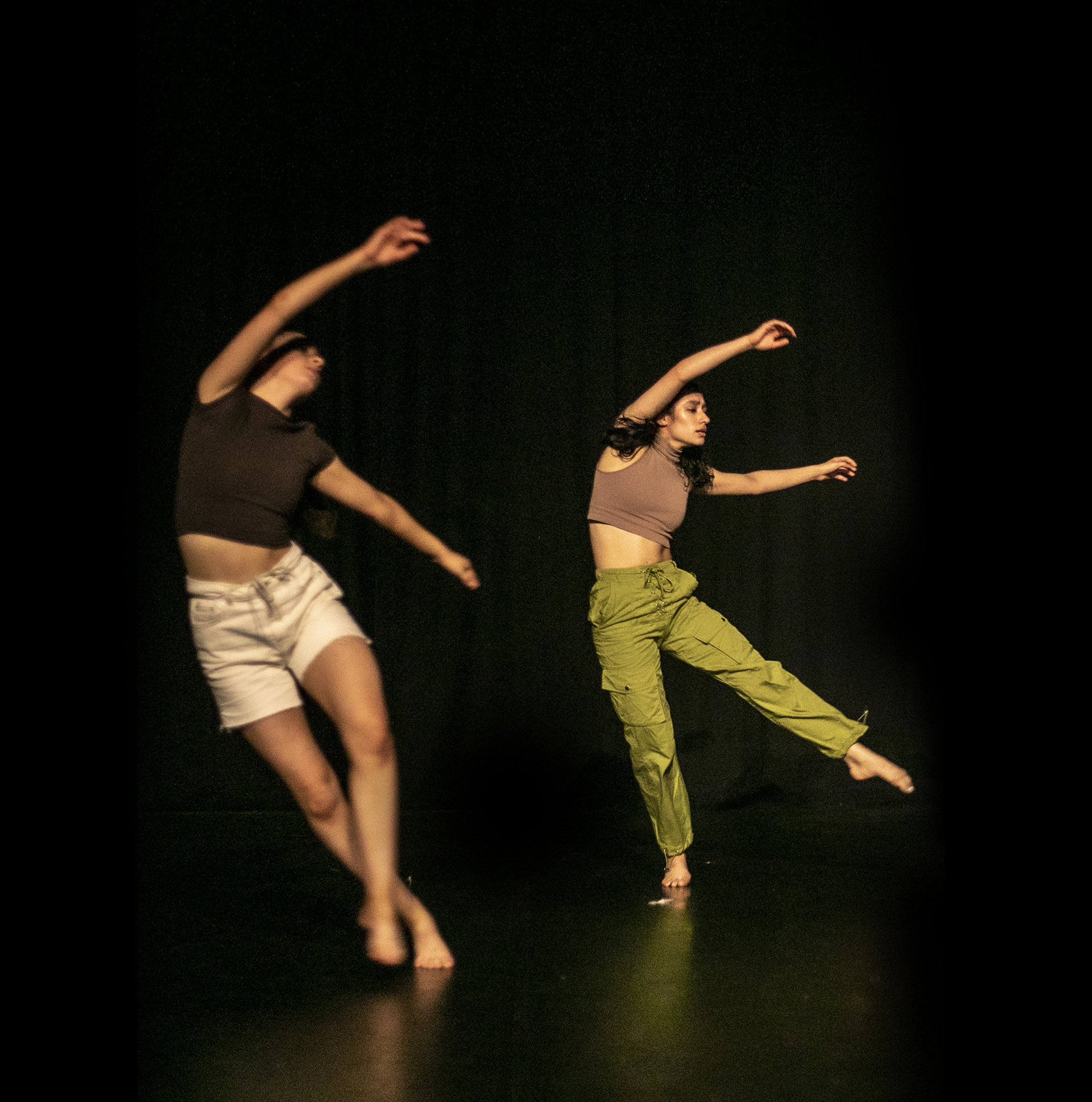 Two female dancers on a black stage with arms in the air feet together leaning to the left. Wearing neutral tones. 