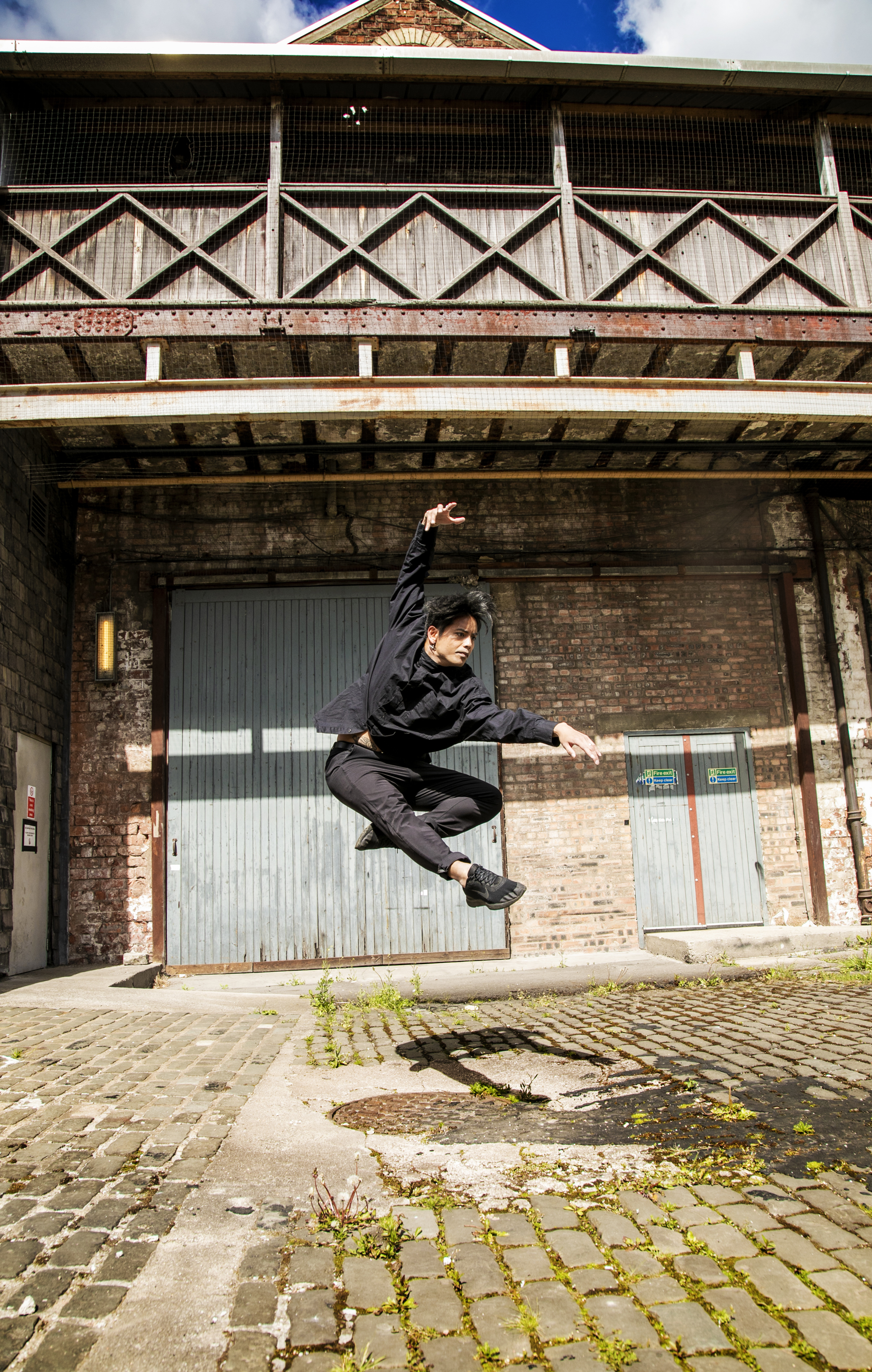 Global majority male dancer jumping high with one leg tucked behind him and the other strecthed in front of him, with one arm stretched above his head and the other out infront of his face angled to the bottom right of the image. Wearing all black in front of industrial building with wooden balcony and trinagle roof.