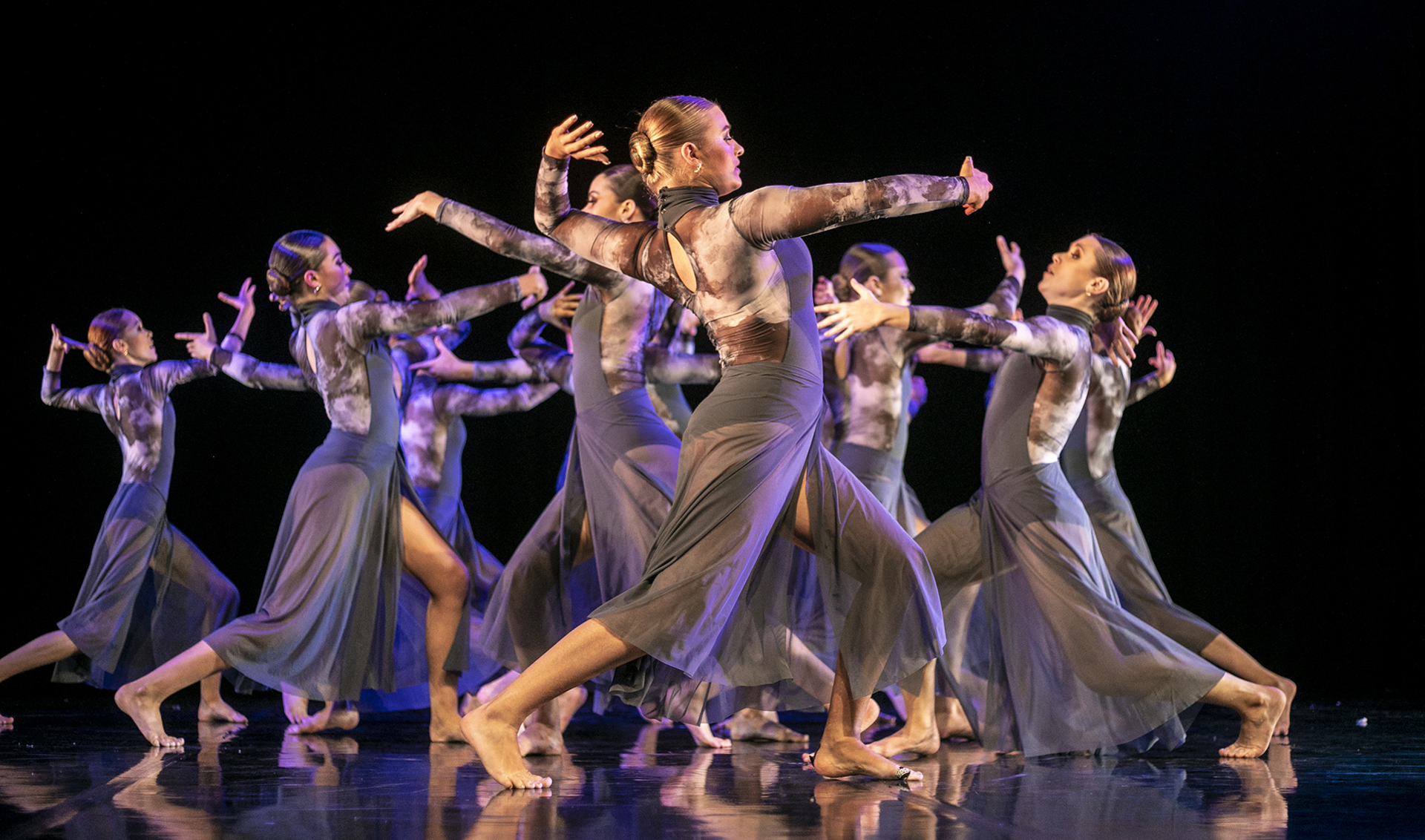 Severn female dancers on a stage wearing long purple jumpsuits with legs in a stride, arched back and arms in front of them scattered around the stage. Severn female dancers on a stage wearing long purple jumpsuits with legs in a stride, arched back and arms in front of them scattered around the stage.