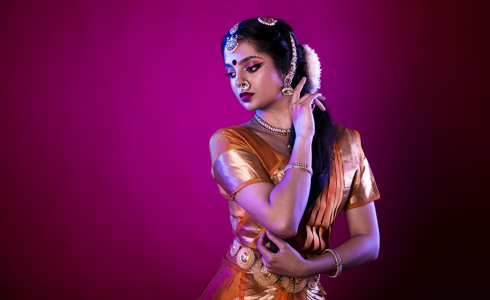 South Asian dancer holding one hand to face and the other towards her elbow on hot pink studio backdrop. Wearing traditional orange dress with gold belt bracelets necklace and head jewlery with gold nose ring.